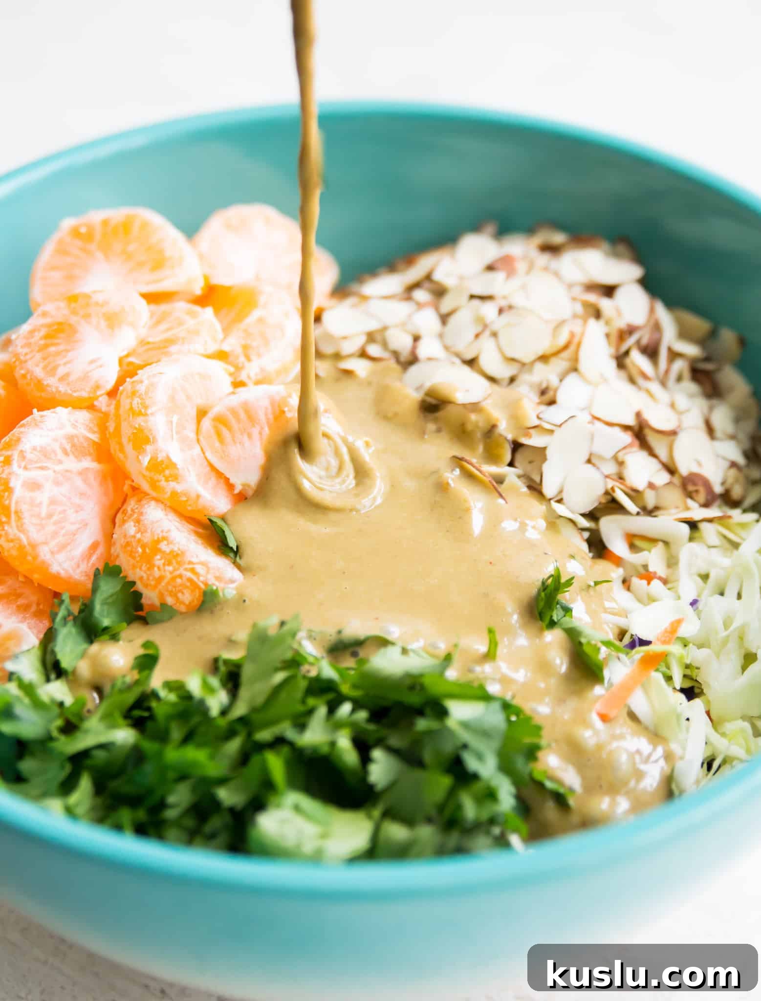 A bowl filled with mandarine orange pieces, chopped cilantro, shredded cabbage and slivered almonds with dressing being poured over the ingredients.