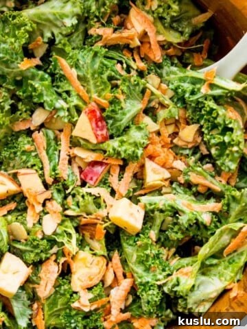 A wooden salad bowl filled with a kale apple salad with a serving spoon in it.