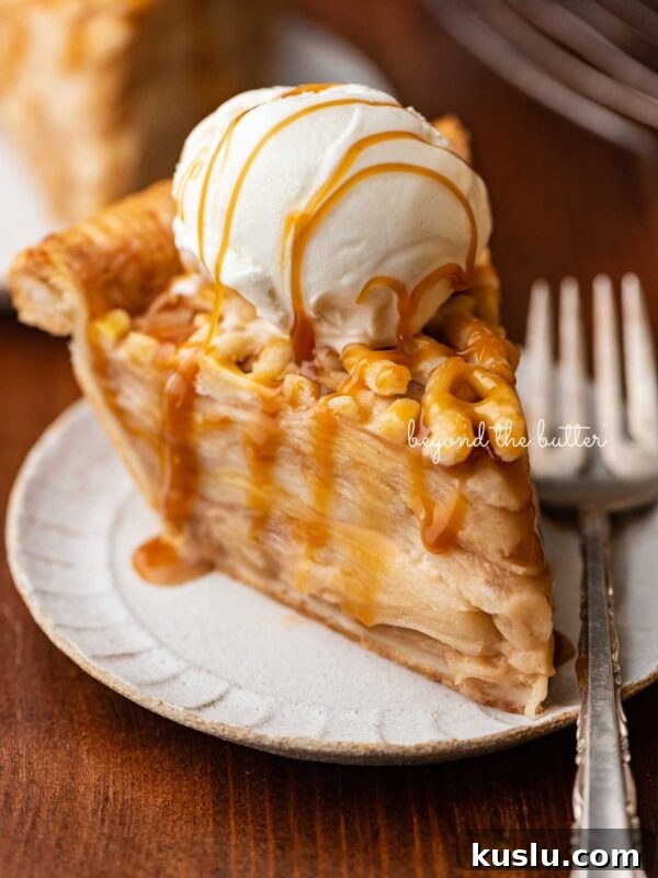 Slice of apple pie drizzled with caramel and topped with a scoop of vanilla bean ice cream on a ceramic dessert plate with a fork placed next it.
