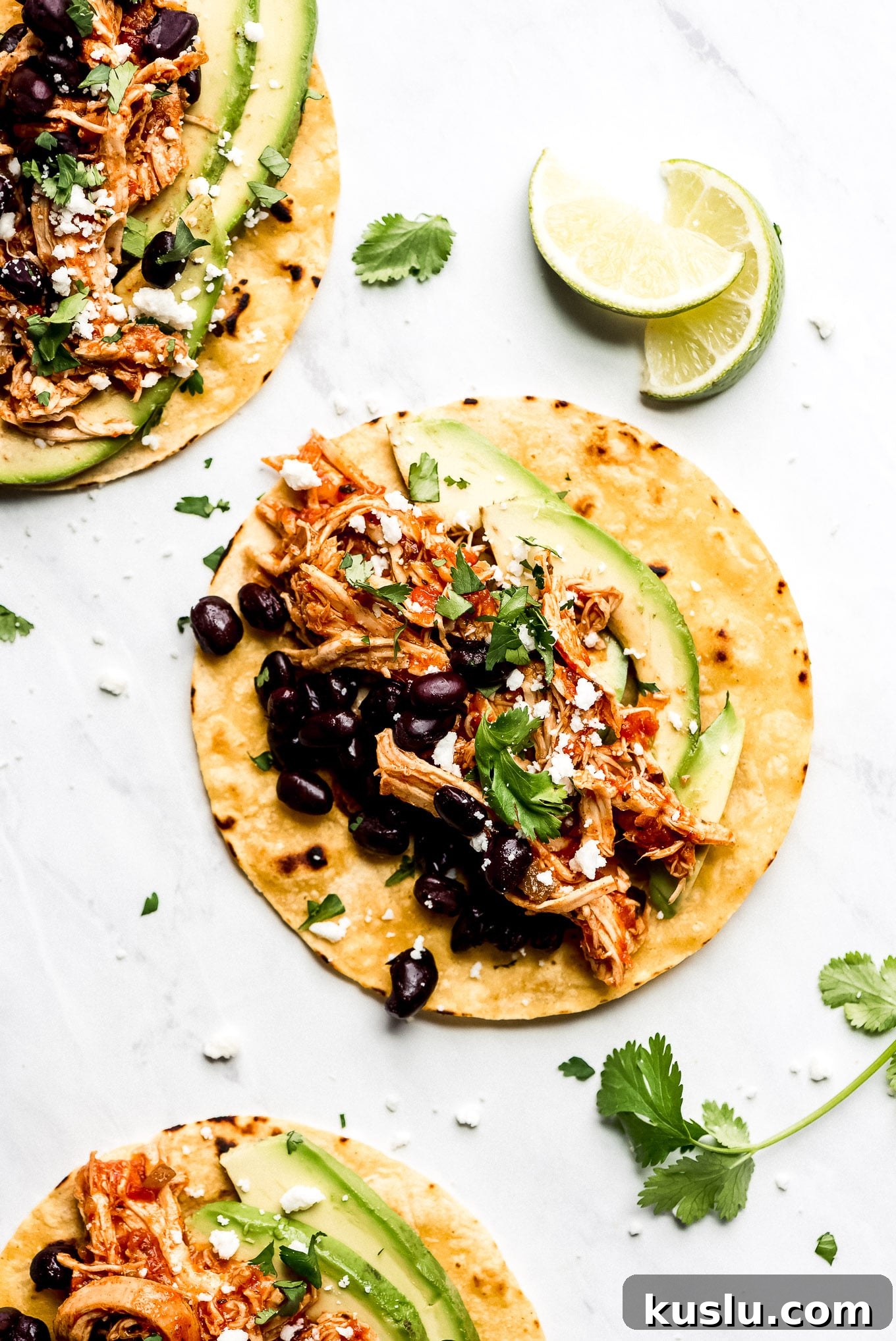 Salsa Chicken Tacos Corn tortillas on a table topped with shredded Salsa Chicken, black beans, avocado slices, cilantro, and Queso Fresco.
