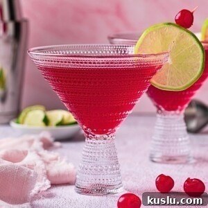 Red pomegranate martini in a clear cocktail glass with a lime wedge and cherry for a garnish.