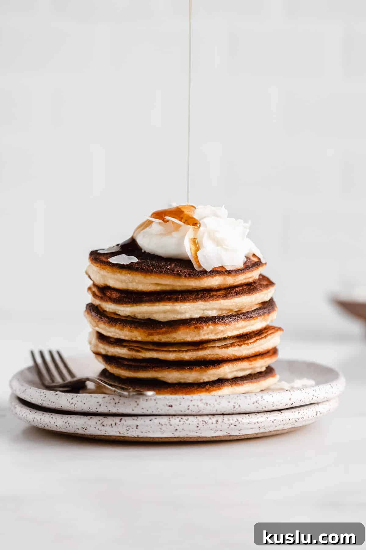 a stack of coconut flour pancakes topped with yogurt and coconut flakes with maple syrup being drizzled on top