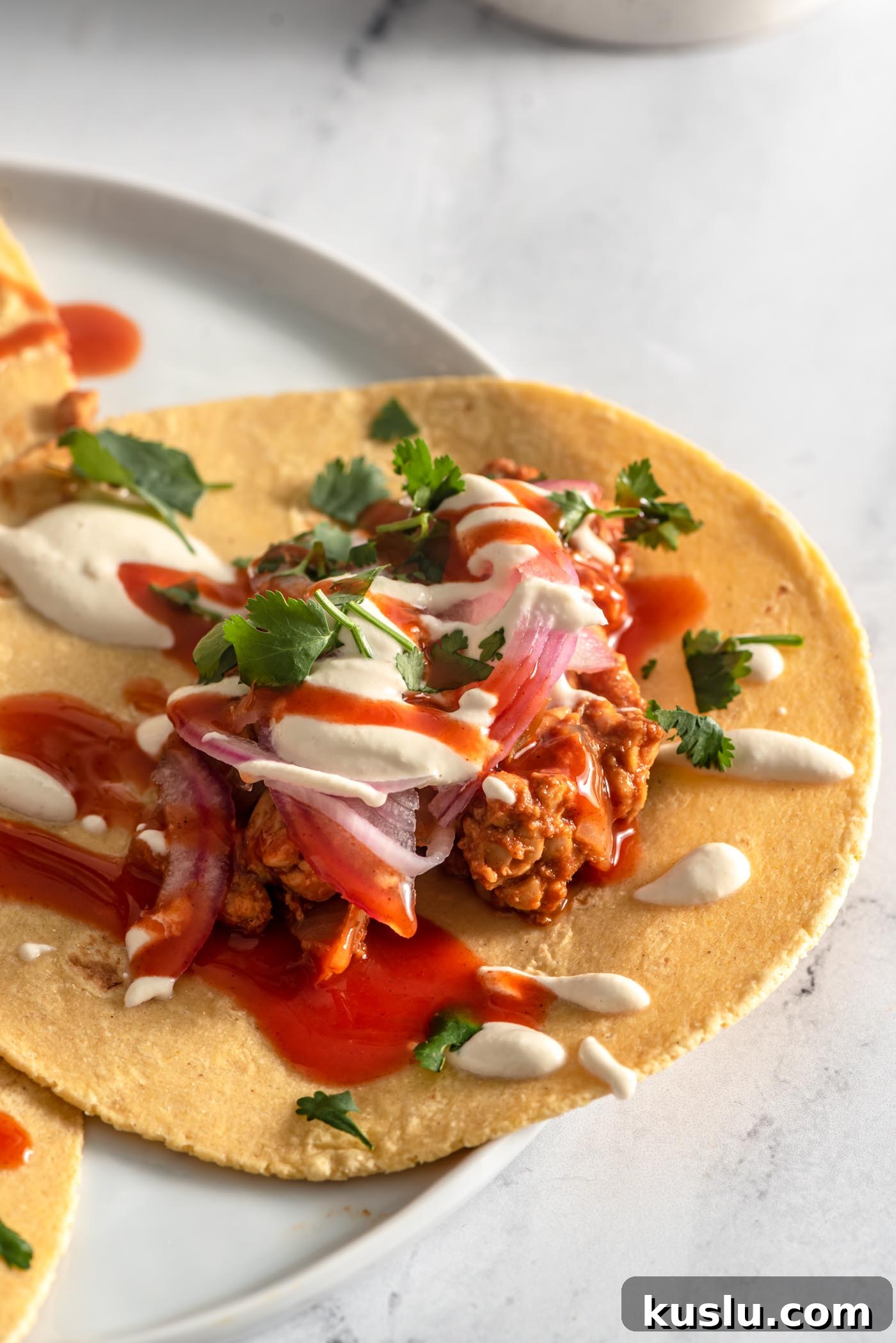 Close up of a tempeh taco topped with pickled red onion, cilantro, cashew cream sauce and hot sauce.