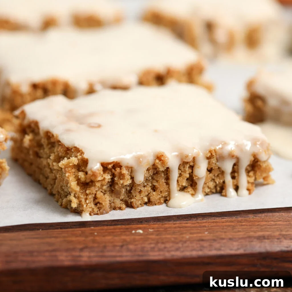 A close-up of a square, frosted Maple Blondie with a bite taken out reveals its moist, crumbly texture. The glaze drips down the side, and more maple blondies appear in the blurred background.