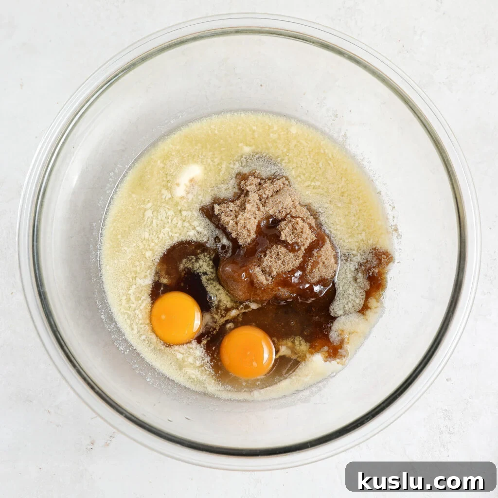 A glass bowl containing two cracked eggs, melted butter, brown sugar, and vanilla extract on a white surface, ready to be mixed for baking delicious Maple Blondies.