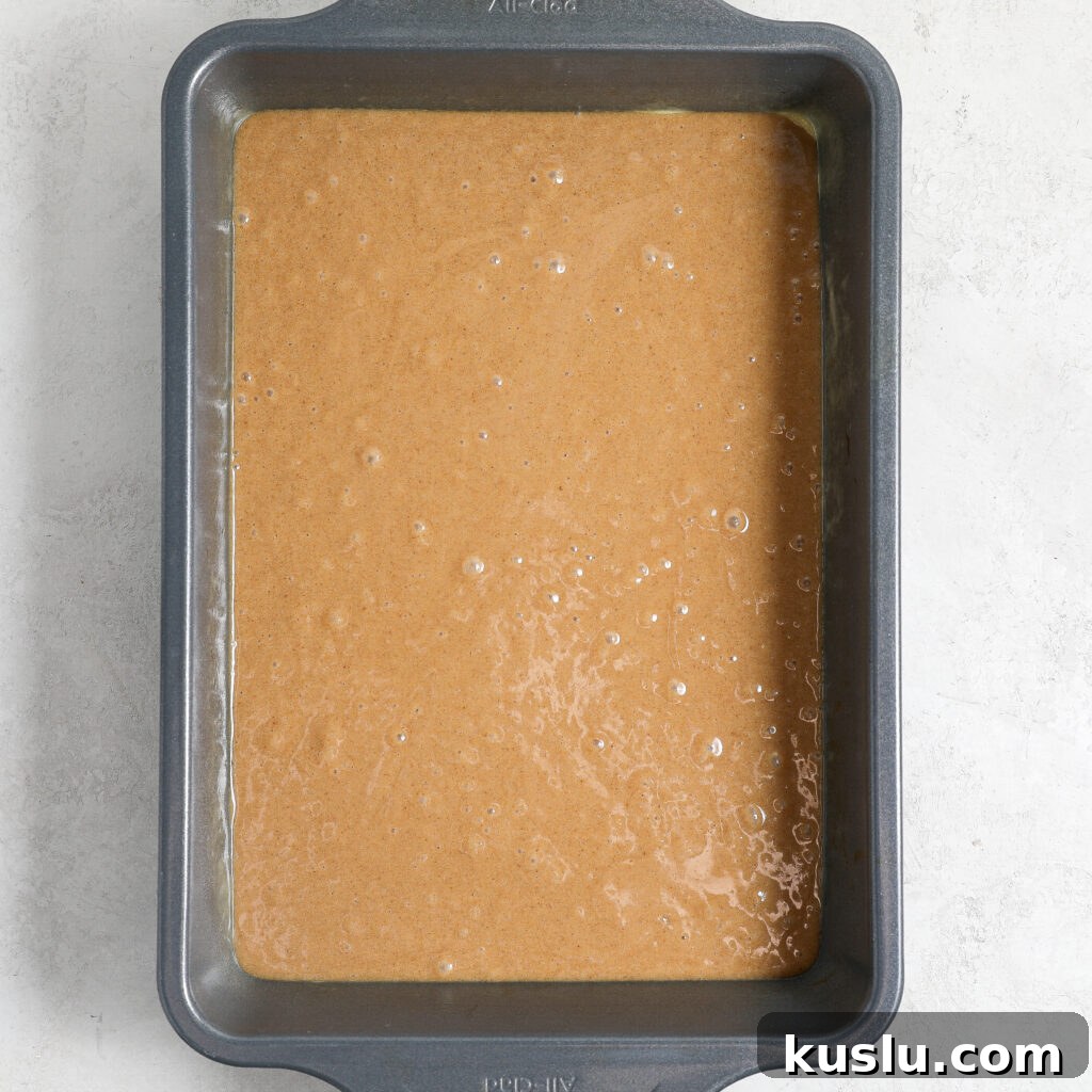 A rectangular metal baking pan filled with smooth, light brown maple blondies batter sits on a light-colored surface. The batter has a few small bubbles rising, ready to be baked into delicious, maple-infused treats.