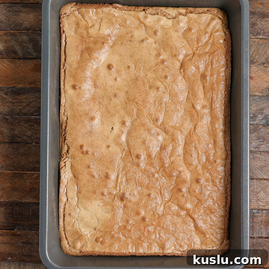 A rectangular baking pan filled with golden-brown maple blondies rests on a rustic wooden surface. The blondies have a slightly cracked top and appear soft and freshly baked.