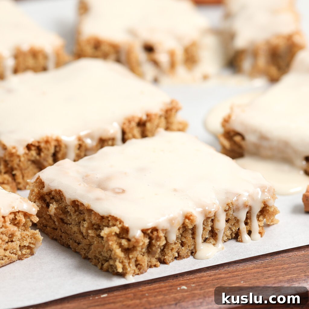 Close-up of several frosted Maple Blondies on parchment paper. The bars have a golden-brown color and thick, creamy glaze drizzling down the sides. They are cut into squares and set on a wooden surface.
