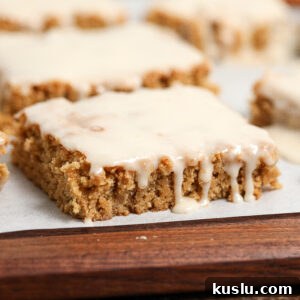 A close-up of a square, frosted Maple Blondie with a bite taken out reveals its moist, crumbly texture. The glaze drips down the side, and more maple blondies appear in the blurred background.