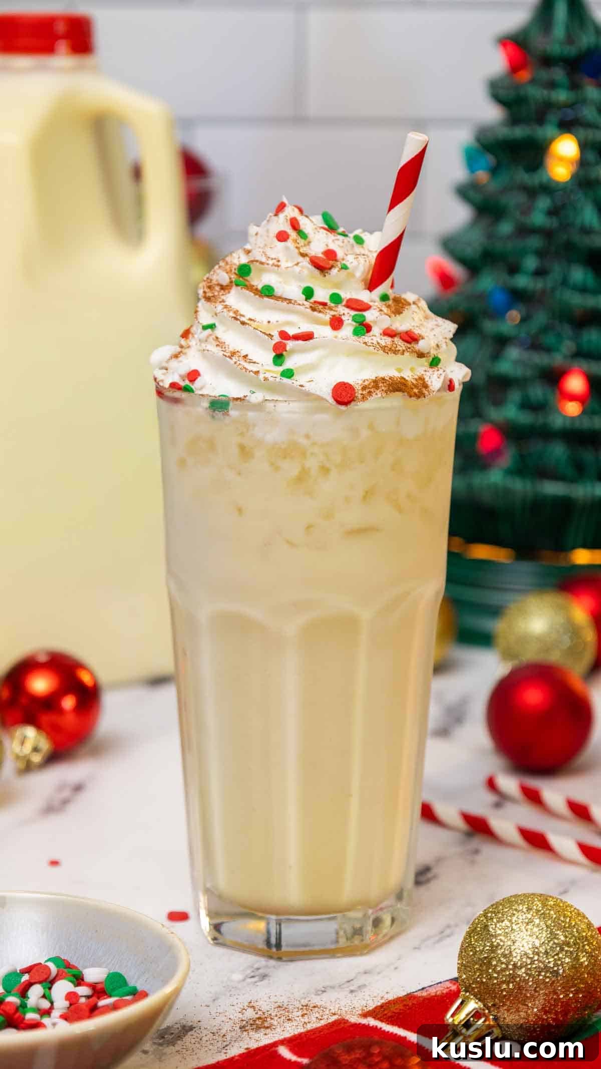 Holiday Eggnog Dirty Soda Recipe — Festive Creamy Cocktail 2 Tall glass of eggnog dirty soda topped with whipped cream, red and green holiday sprinkles, and a dusting of cinnamon, served with a red-striped straw. The festive background includes Christmas ornaments, a ceramic Christmas tree, and a carton of eggnog.