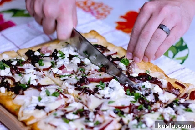 cutting pizza on a cutting board featuring pear and goat cheese flatbread topped with caramelized onions, ham and cheddar