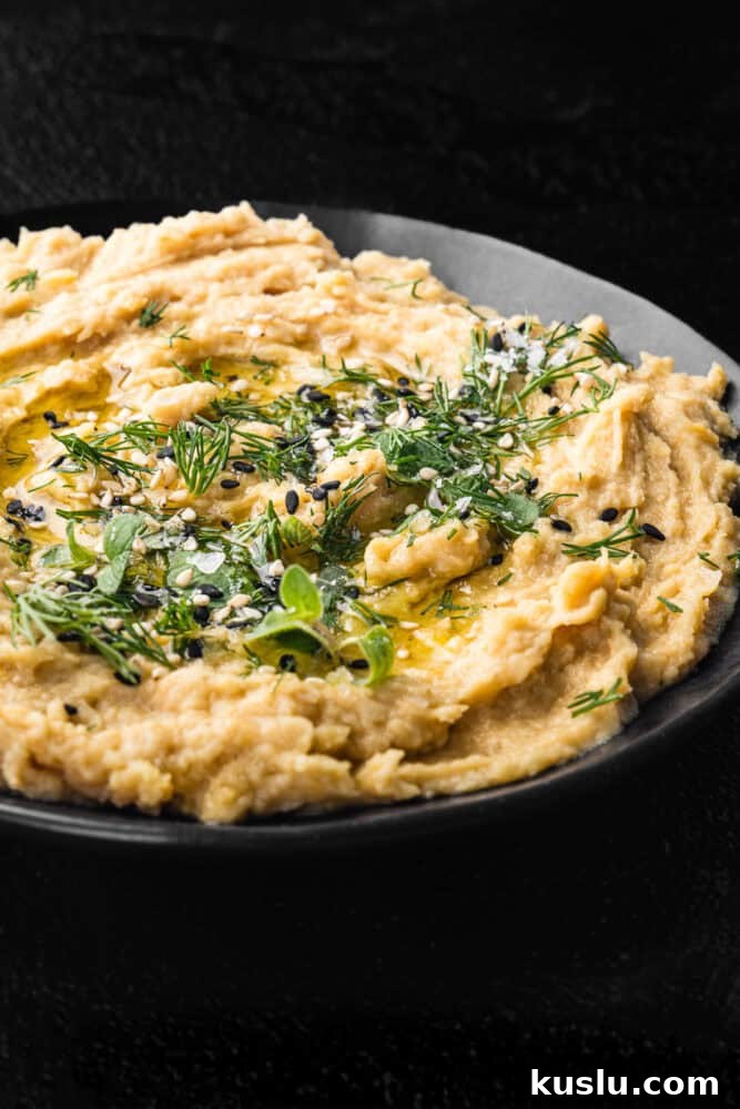 Hummus without tahini topped with fresh dill, sesame seeds, olive oil, and flaky salt in a black bowl on a dark background, close-up angled shot