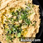 Overhead shot of hummus without tahini in a black bowl, topped with fresh dill, sesame seeds, olive oil, and flaky sea salt.