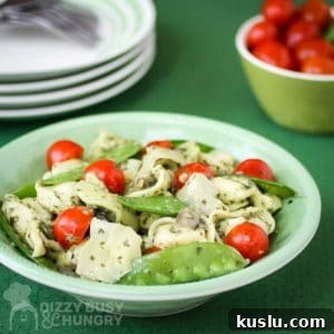 Pesto Tortellini with Artichokes and Snow Peas Recipe 9 Side view of pesto tortellini in a green bowl with a bowl of grape tomatoes and plates in the background.