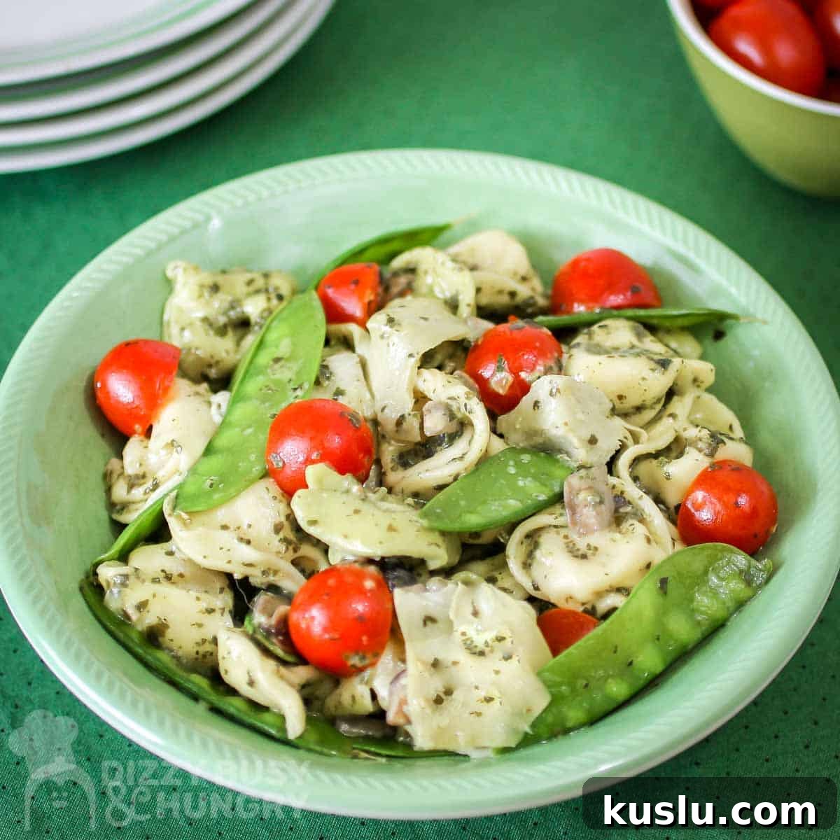 Pesto Tortellini with Artichokes and Snow Peas Recipe 5 Overhead shot of pesto tortellini in a green bowl with a bowl of grape tomatoes and plates in the background.