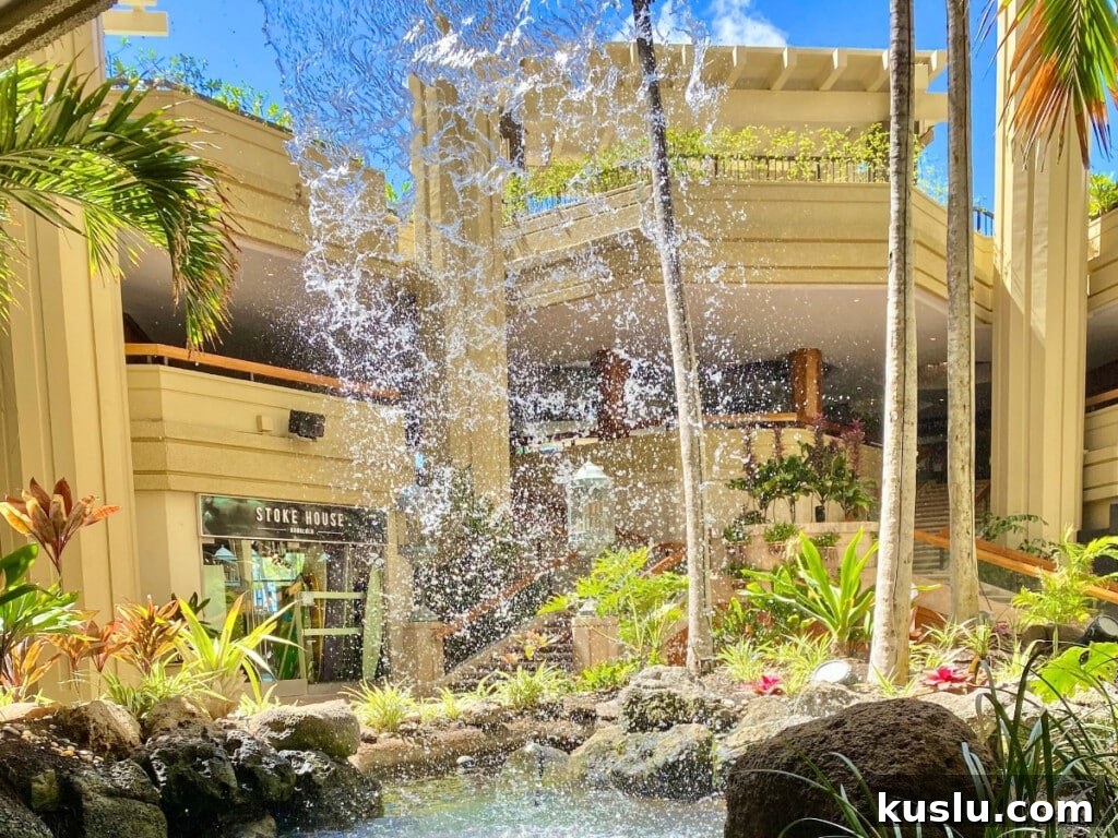 Scenic view of the Pualeilani Atrium Shops at Hyatt Regency Waikiki Beach Resort, featuring a vibrant tropical courtyard with a fountain, lush greenery, and open-air architecture.