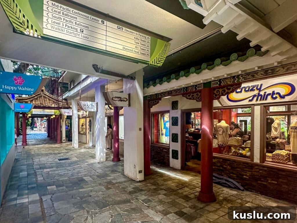 Pathway at Hilton Hawaiian Village shopping area in Waikīkī, Hawaiʻi featuring Crazy Shirts and other boutique stores, with tiled stone flooring, traditional architectural accents, and tropical signage.