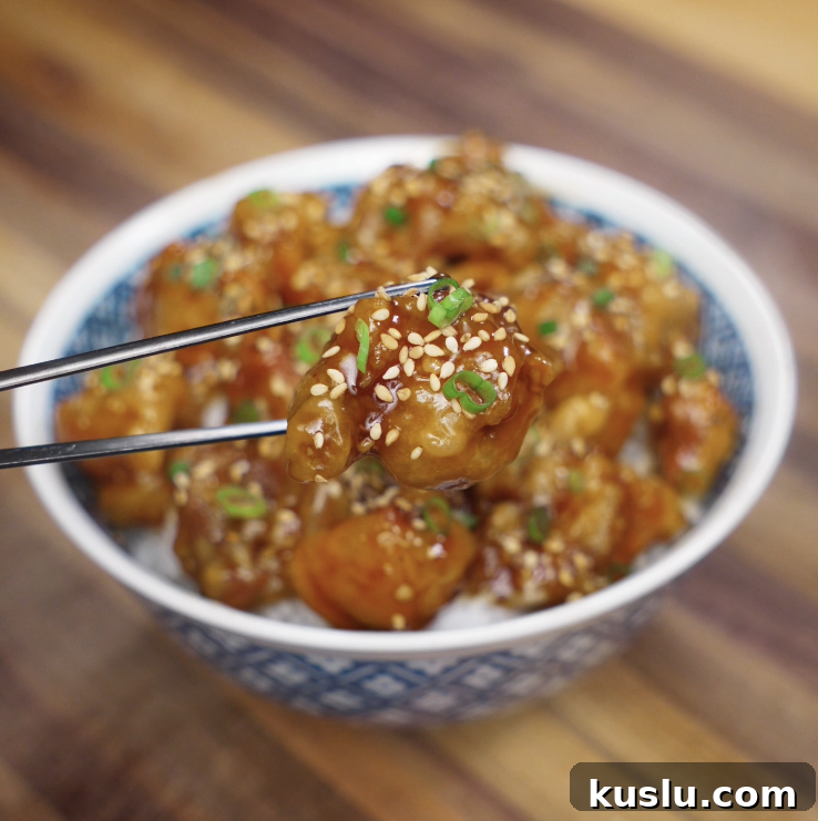 Crispy Korean Fried Chicken Recipe: How to Make Dakgangjeong 3 A glossy, golden piece of Korean fried chicken is lifted from the bowl with chopsticks, showing the sticky glaze.