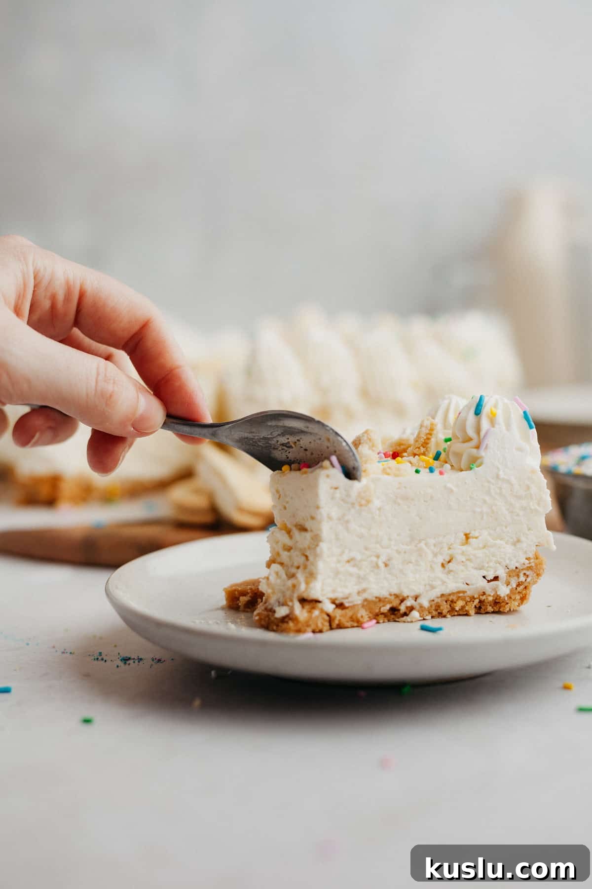 A slice of no bake cheesecake covered in rainbow sprinkles on a small beige plate. A hand is holding a small silver spoon and is cutting into the slice