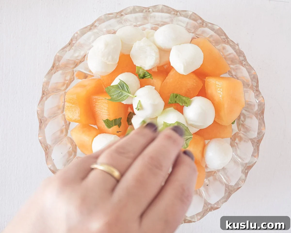 A glass bowl with cantaloupe melon cubes, mozzarella balls and basil.