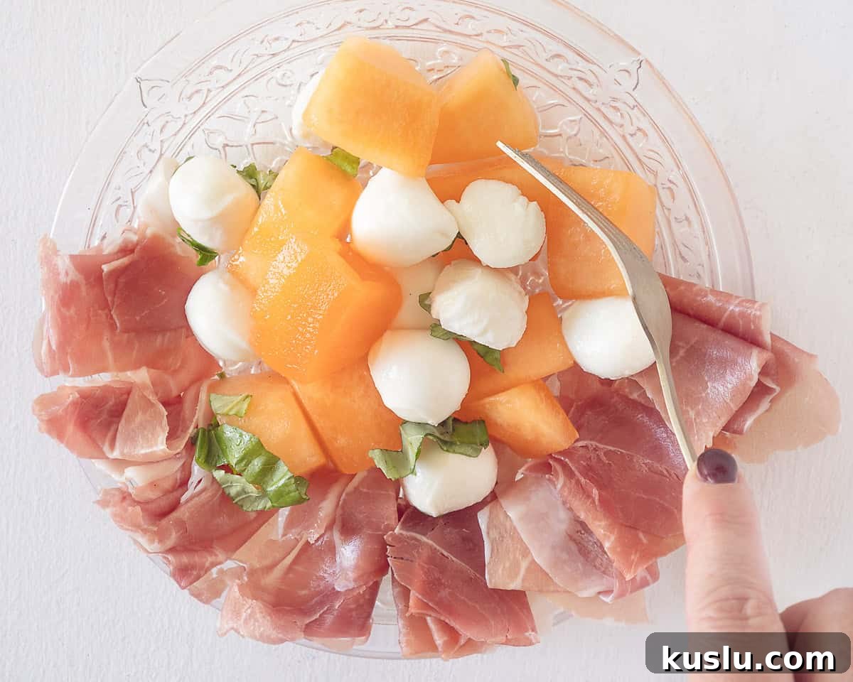 A glass plate with parmaham, mozzarella, melon, basil and dressing. A fork mixing everything.