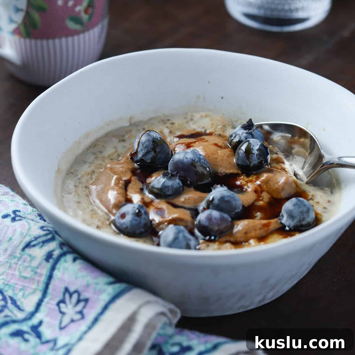 Blueberry Oat & Chia Porridge with Almond Butter (Vegan) 2 photo of a white bowl of vegan oat and chia seed porridge with blueberries and coconut almond butter on top, all on a dark wood table with a napkin to the side and cup of coffee in the background.