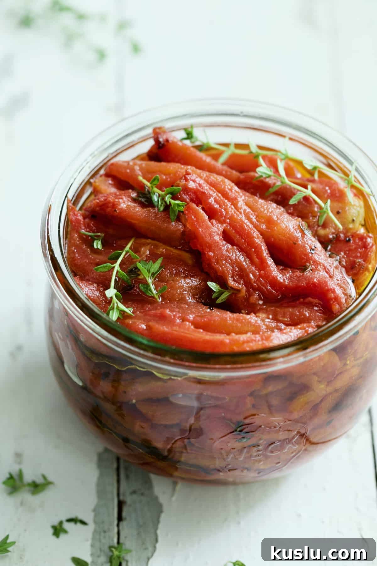 Tomato Confit in glass jar garnished with fresh thyme sprigs on white distressed background.