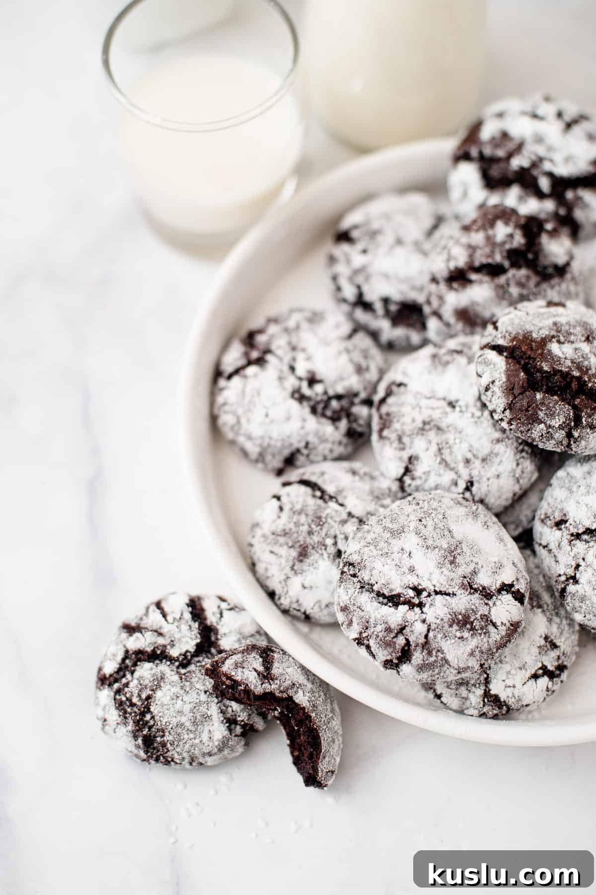 Chewy Chocolate Crinkle Cookies Recipe for Perfect Cracks 3 chocolate crinkles served on a white round plate with two cookies sitting next to the plate on a marble countertop.