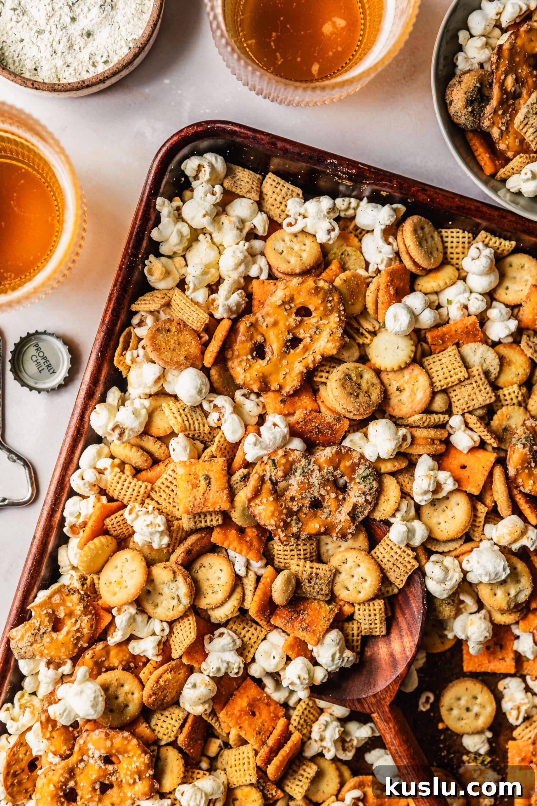 Spicy Ranch Snack Mix Recipe: Zesty Party Mix Everyone Loves 2 A pan of spicy ranch snack mix on a pink counter next to beer, a grey bowl of mix and a brown bowl of ranch seasoning.