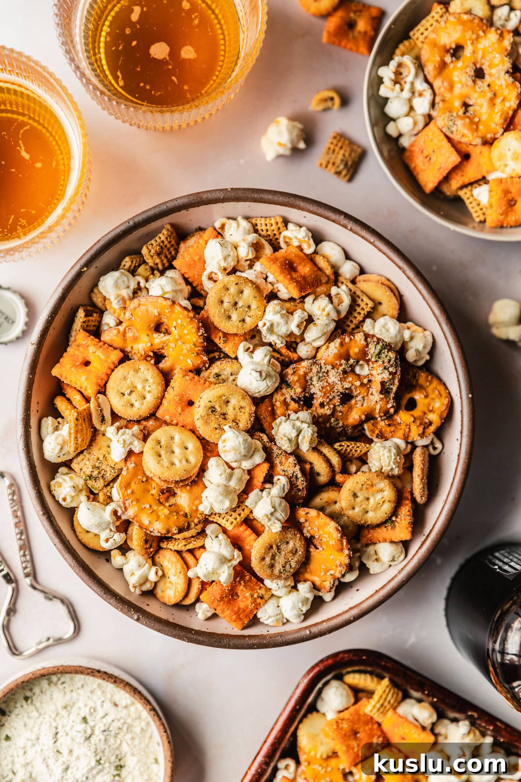 Spicy Ranch Snack Mix Recipe: Zesty Party Mix Everyone Loves 11 A white bowl of spicy ranch snack mix on a pink counter next to a grey bowl of snacks, glasses of beer, and a brown bowl of ranch seasoning.