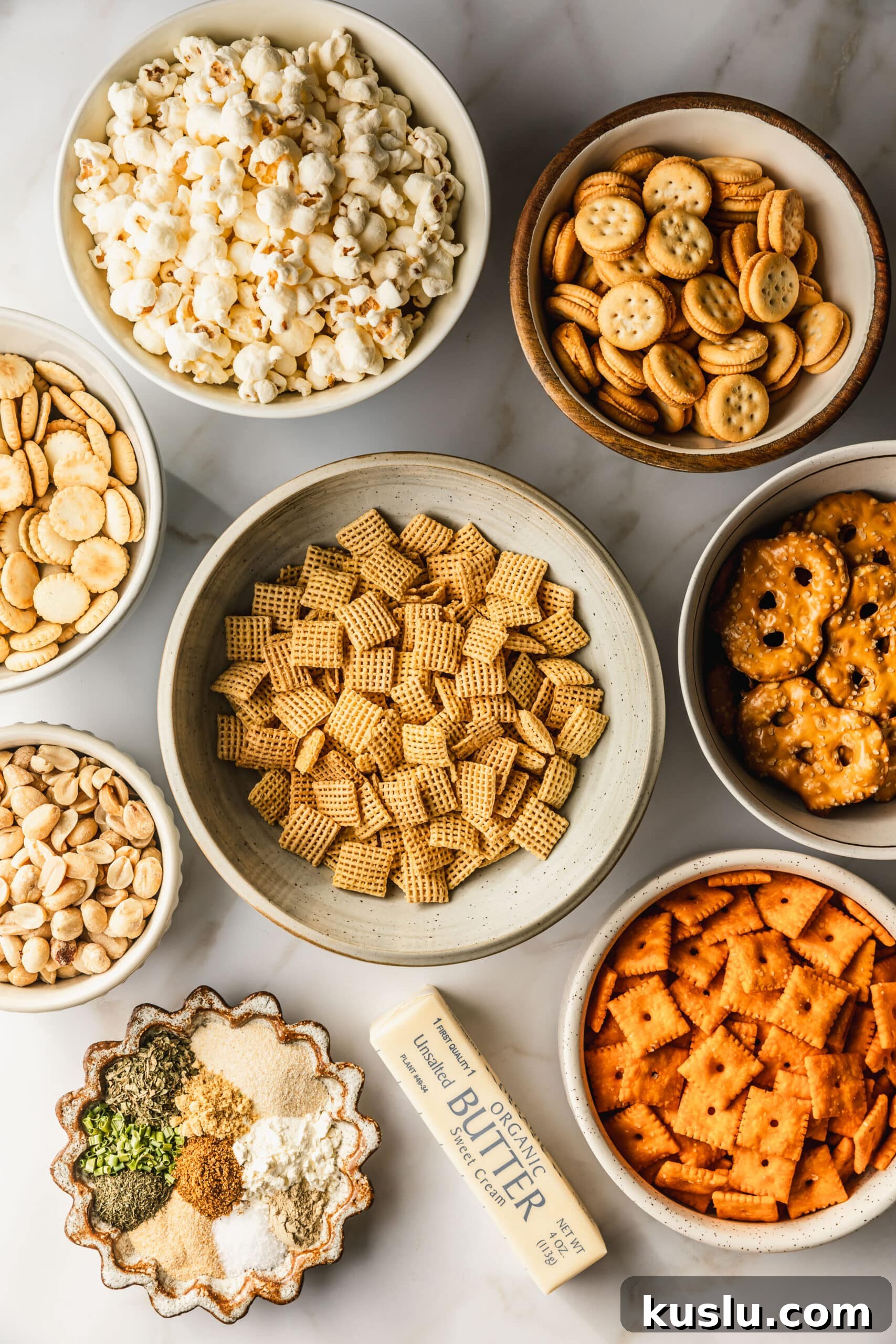 Spicy Ranch Snack Mix Recipe: Zesty Party Mix Everyone Loves 3 White and brown bowls of cereal, pretzels, Cheez Its, peanuts, spices, oyster crackers, sandwich crackers, popcorn, and butter on a white counter.