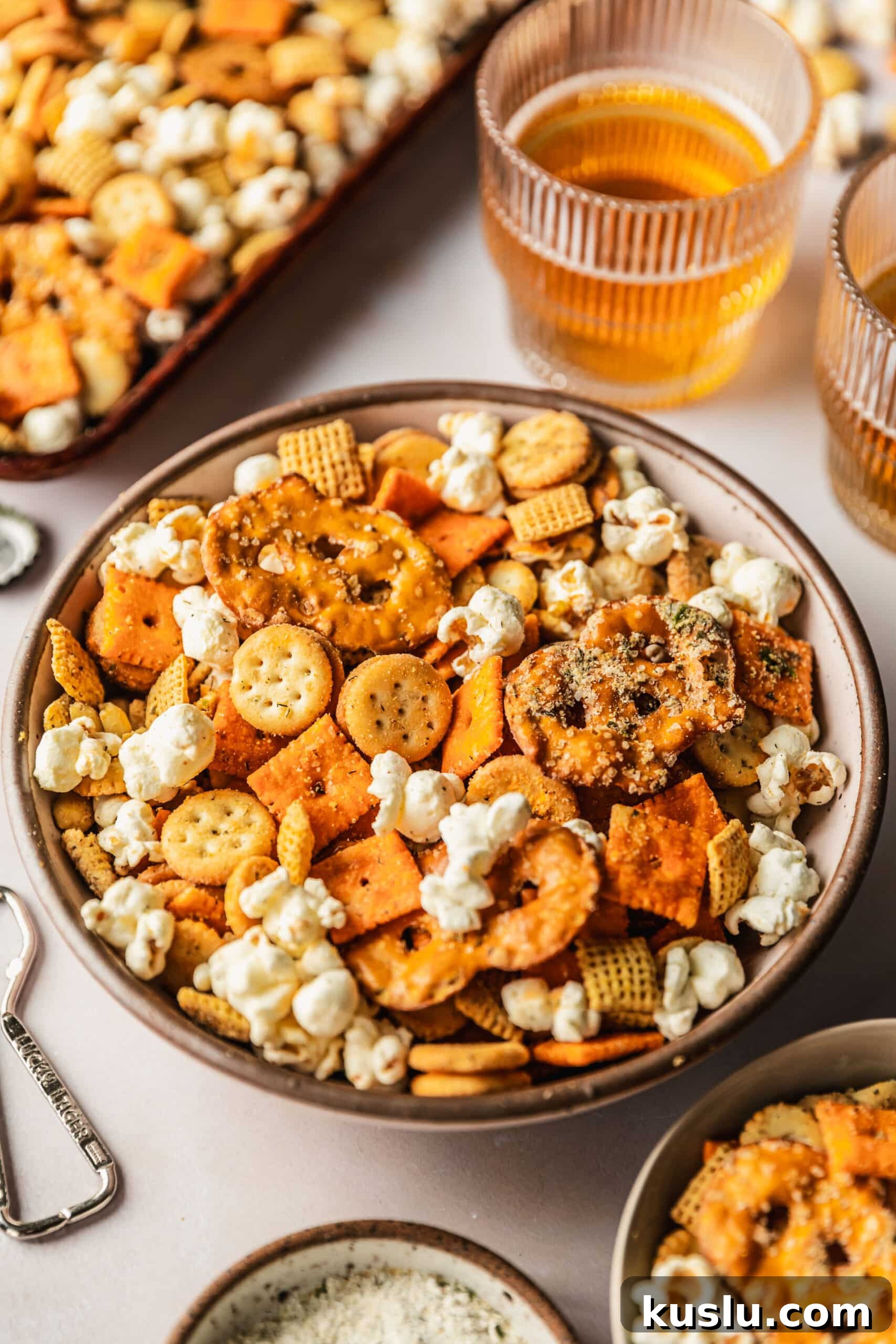 Spicy Ranch Snack Mix Recipe: Zesty Party Mix Everyone Loves 9 A pink bowl of spicy ranch snack mix on a pink counter next to beer, a pan of snack mix, and a bottle opener.