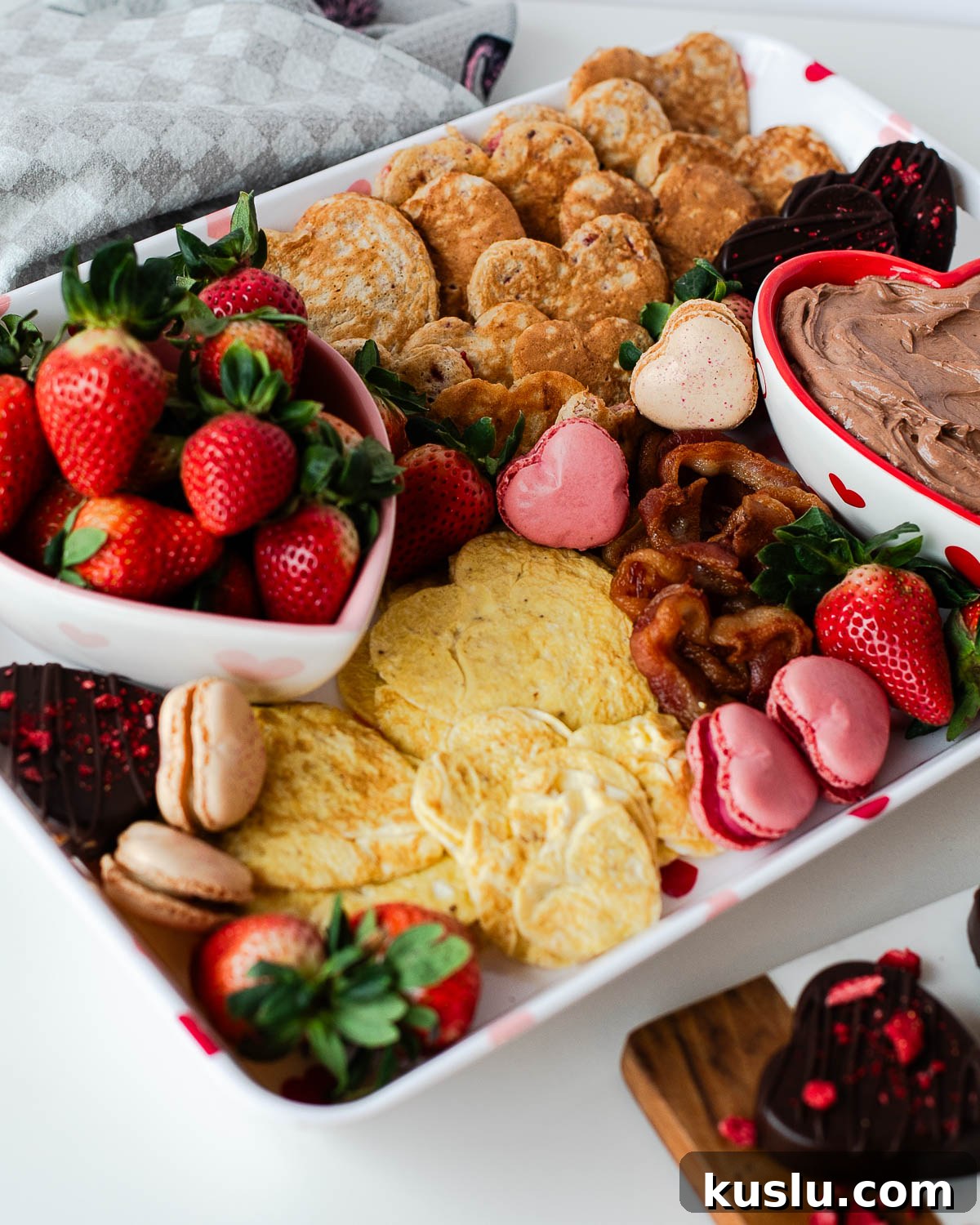 Heart-shaped eggs, crispy bacon, and pink heart-shaped macarons on a Valentine breakfast board.