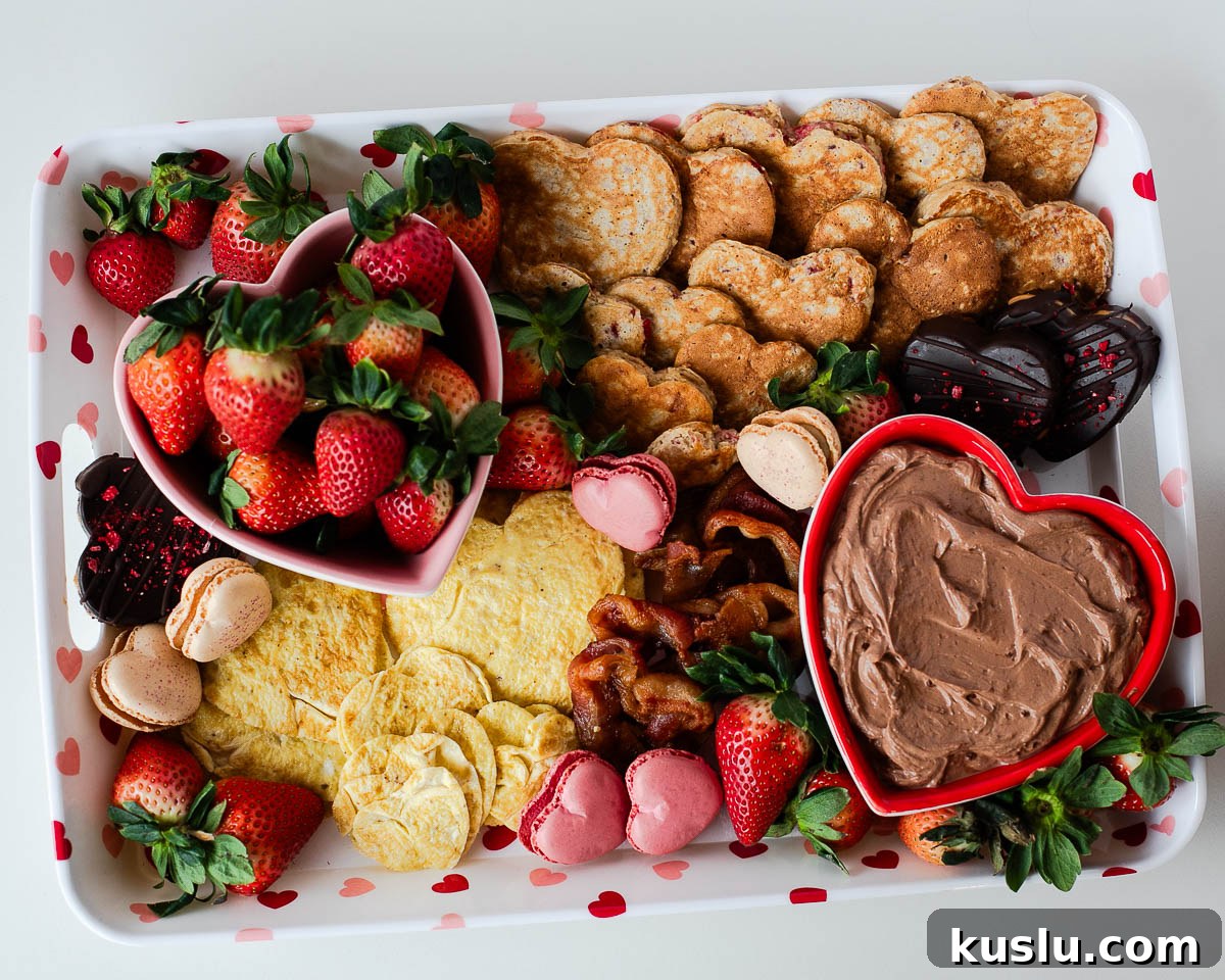 Overhead view of a Valentine breakfast board with heart-shaped pancakes, heart-shaped eggs, bacon, strawberries, macarons, and chocolate yogurt dip.