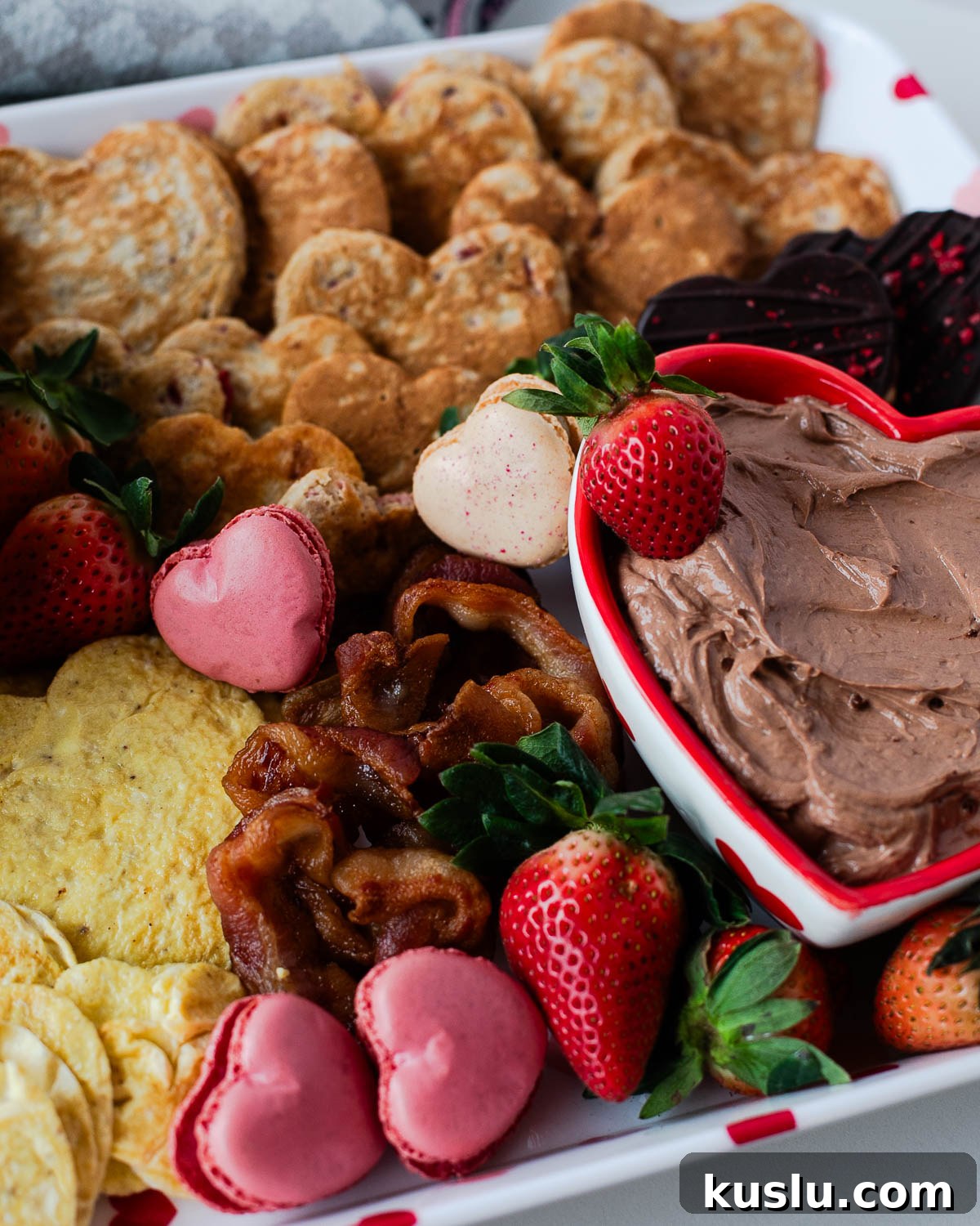 Heart-shaped eggs, crispy bacon, and pink heart-shaped macarons on a Valentine breakfast board.