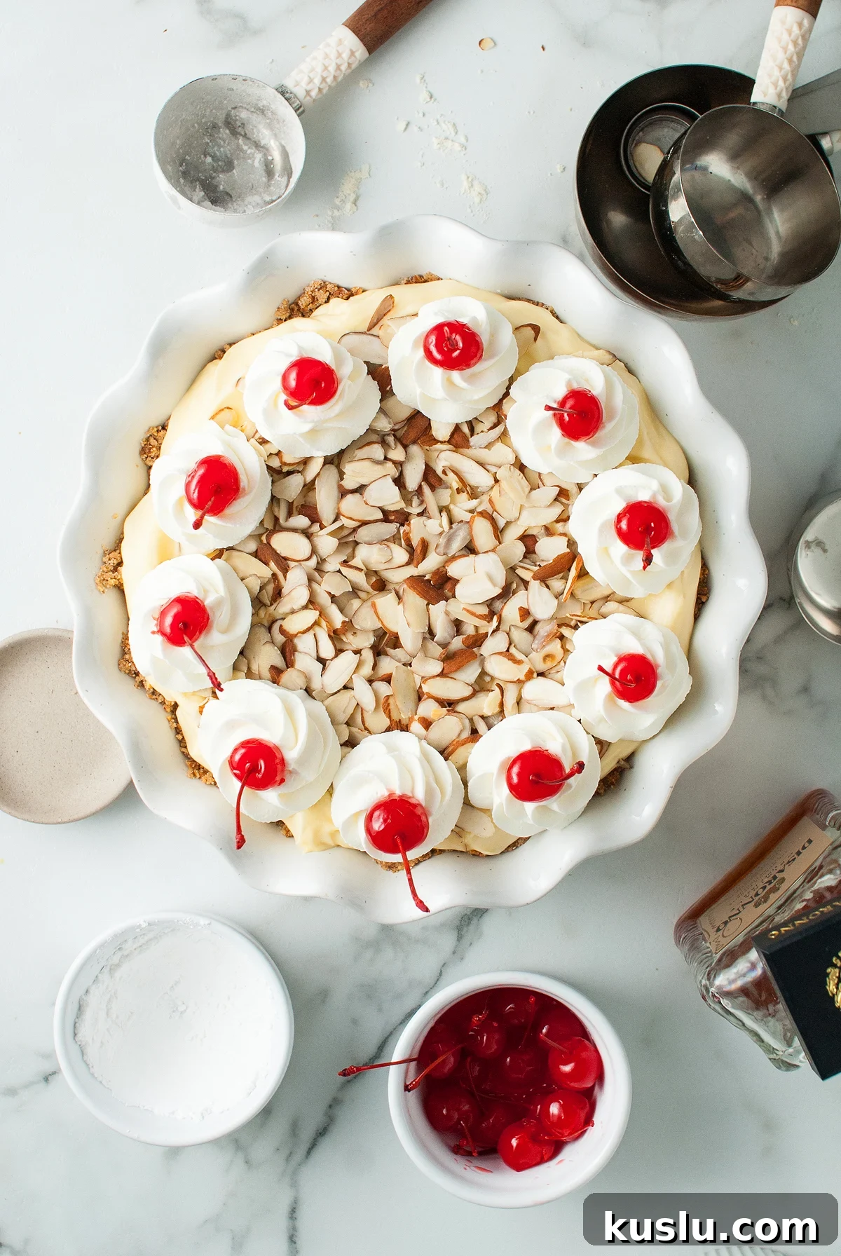 Decadent Amaretto Cream Pie Recipe with Almond Liqueur Filling 11 An Almond amaretto pie topped with whipped cream, cherries, and sliced almonds sits on a marble counter with baking tools.