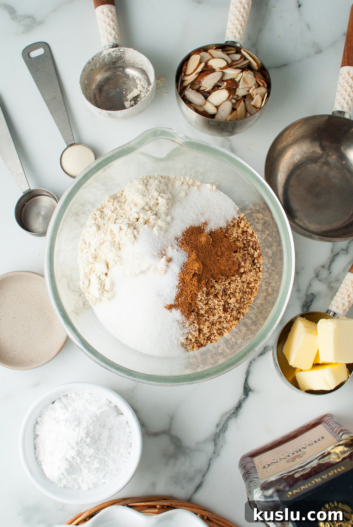 Decadent Amaretto Cream Pie Recipe with Almond Liqueur Filling 14 Bowl with flour, sugar, cinnamon, and nuts surrounded by baking ingredients and utensils on a marble counter—perfect for preparing an Italian inspired dessert like Amaretto Cream Pie.