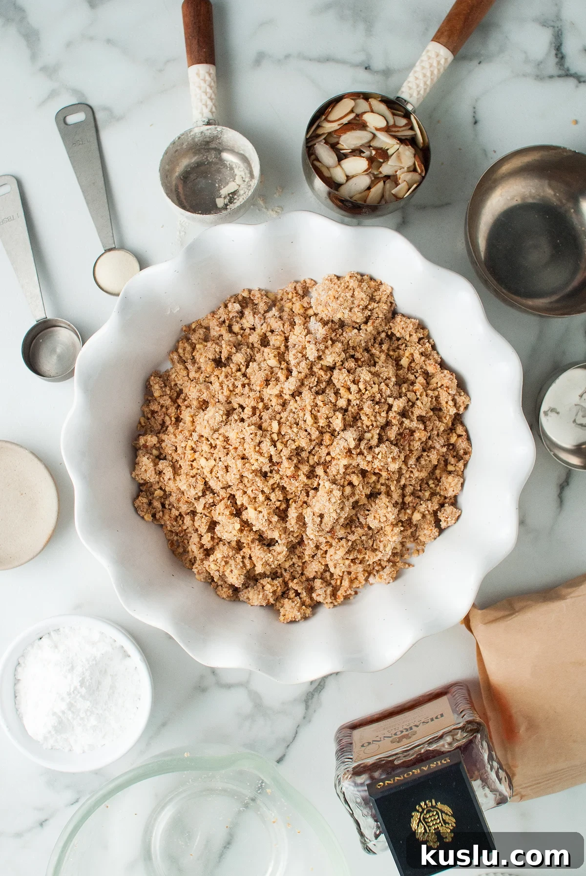 Decadent Amaretto Cream Pie Recipe with Almond Liqueur Filling 15 A pie dish with crumbly topping, surrounded by baking tools, sliced almonds, and ingredients on a marble counter—perfect for creating an Italian inspired dessert like almond amaretto pie.