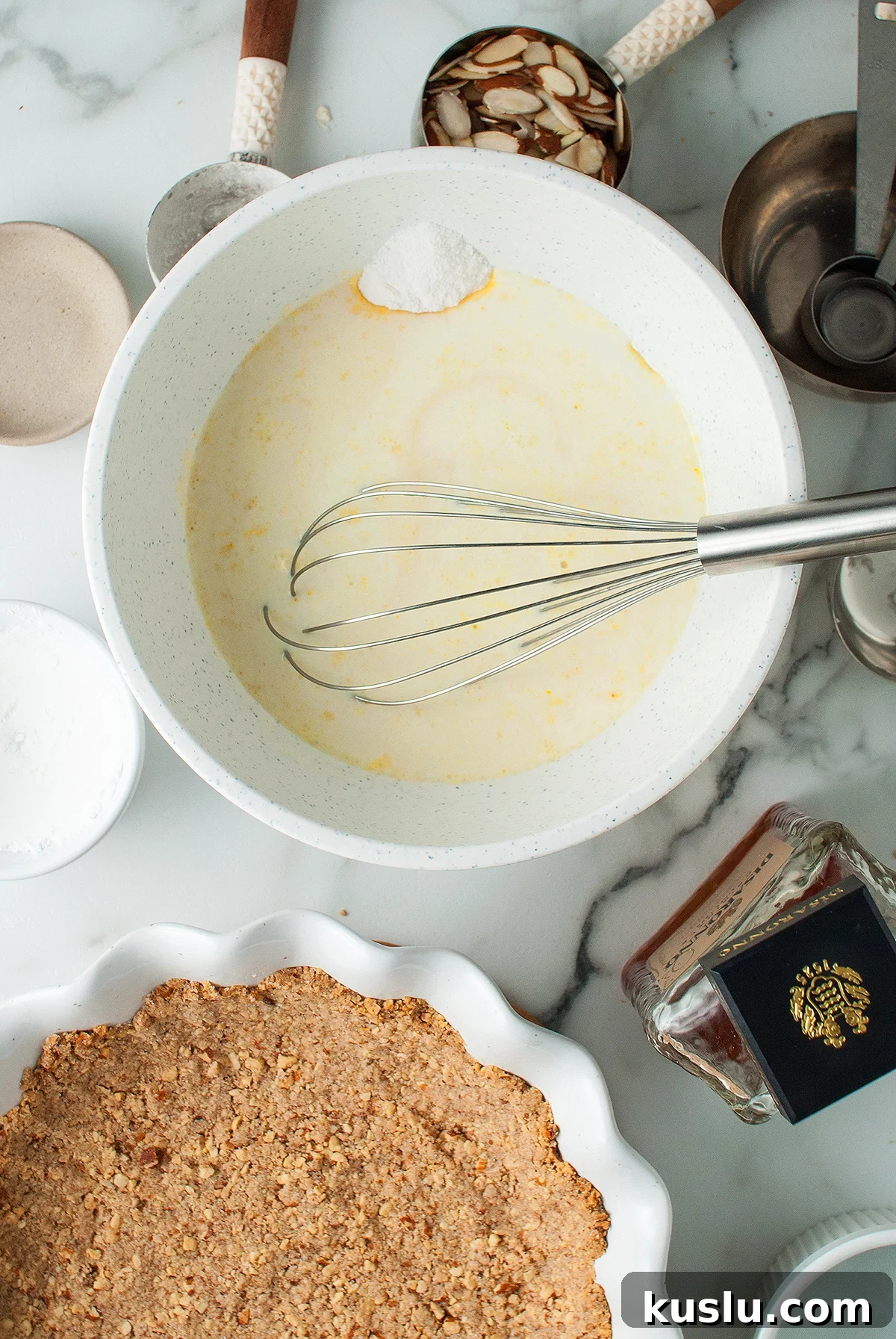 Decadent Amaretto Cream Pie Recipe with Almond Liqueur Filling 16 A bowl with batter and a whisk sits next to an almond crust, cinnamon, and baking ingredients on a countertop—perfect for preparing a delicious almond amaretto pie.
