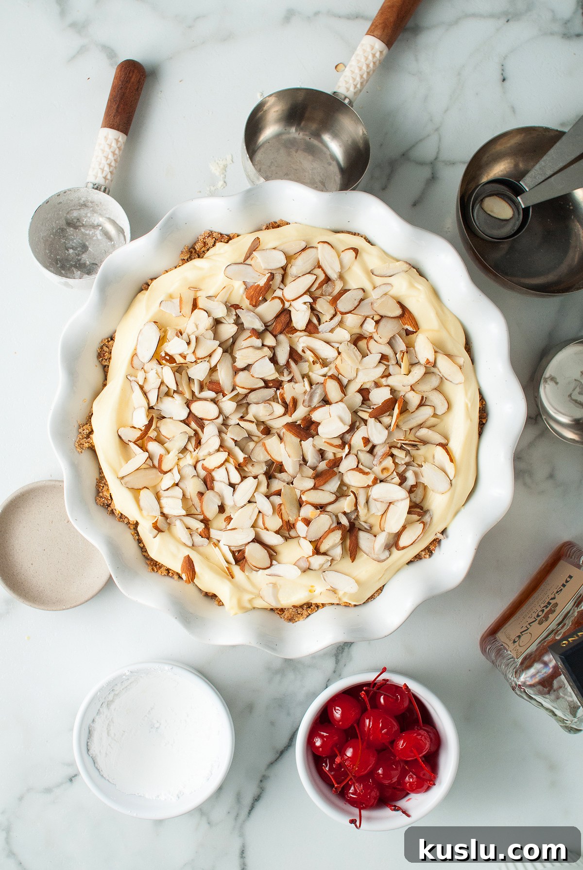Decadent Amaretto Cream Pie Recipe with Almond Liqueur Filling 18 Unbaked Almond amaretto pie topped with sliced almonds, surrounded by cherries, measuring cups, and baking ingredients—an Italian inspired dessert ready to be baked.