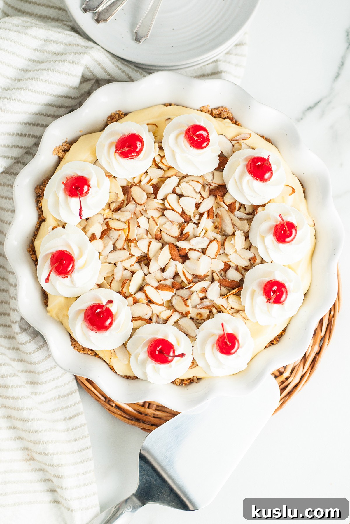 Decadent Amaretto Cream Pie Recipe with Almond Liqueur Filling 7 An Italian inspired dessert, this Amaretto Cream Pie features an almond crust and is topped with whipped cream, cherries, and sliced almonds in a white pie dish on a striped cloth.