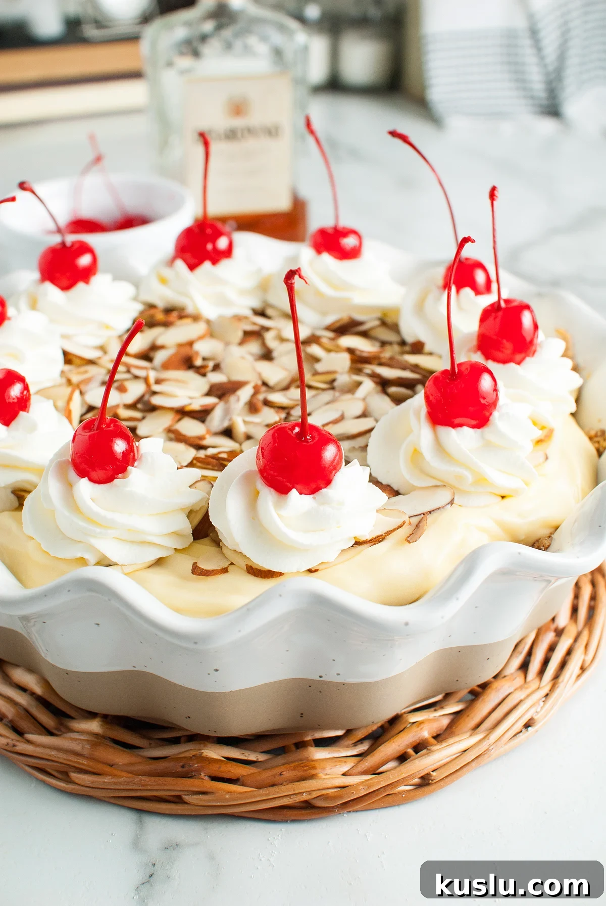 Decadent Amaretto Cream Pie Recipe with Almond Liqueur Filling 9 Creamy Amaretto Cream Pie topped with whipped cream, cherries, and sliced almonds in a white dish on a wicker tray, featuring a rich almond crust for the perfect dessert with amaretto liqueur.