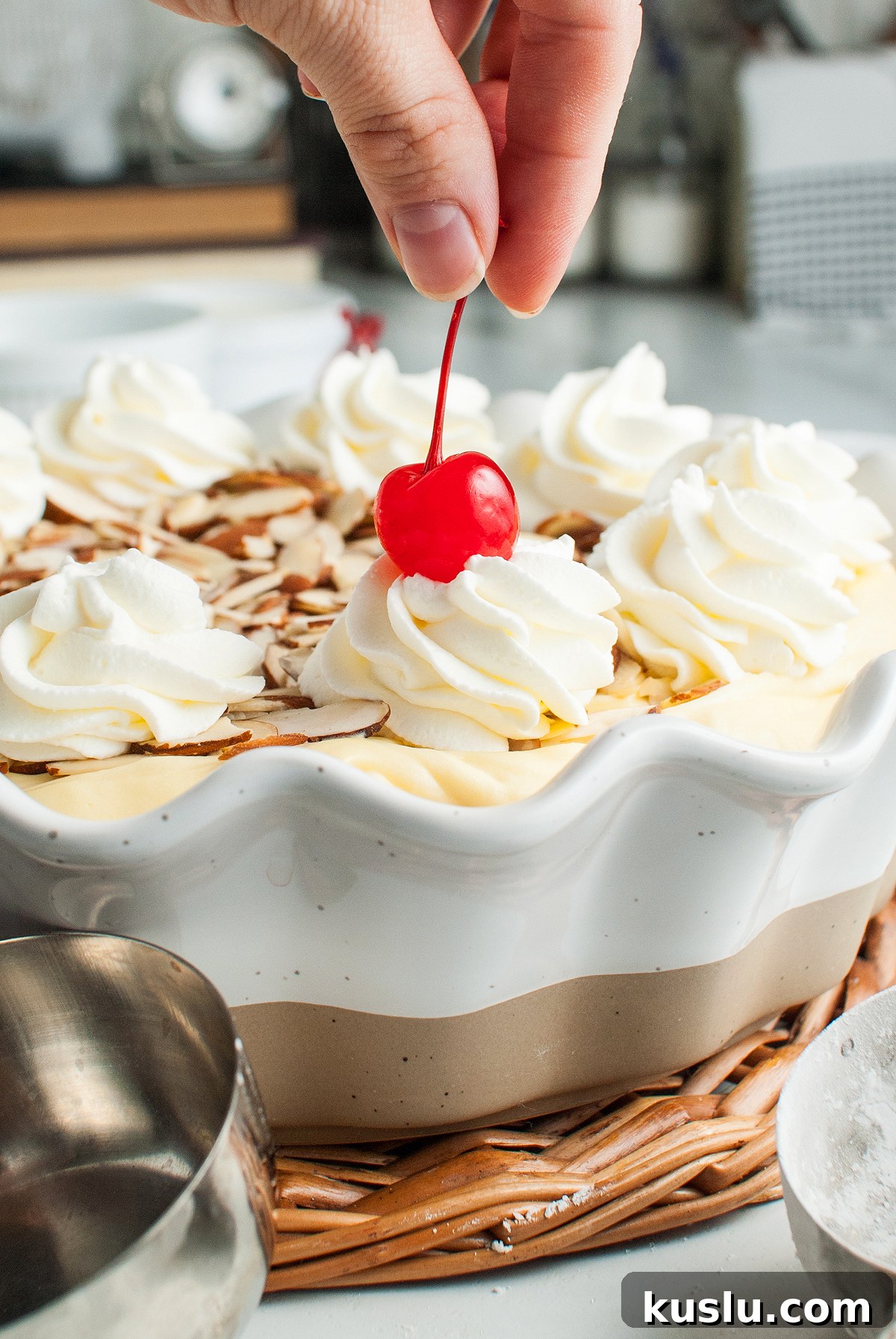 Decadent Amaretto Cream Pie Recipe with Almond Liqueur Filling 10 A hand places a cherry on top of whipped cream on an Italian inspired dessert pie with almond slices and Almond Crust in a white dish.