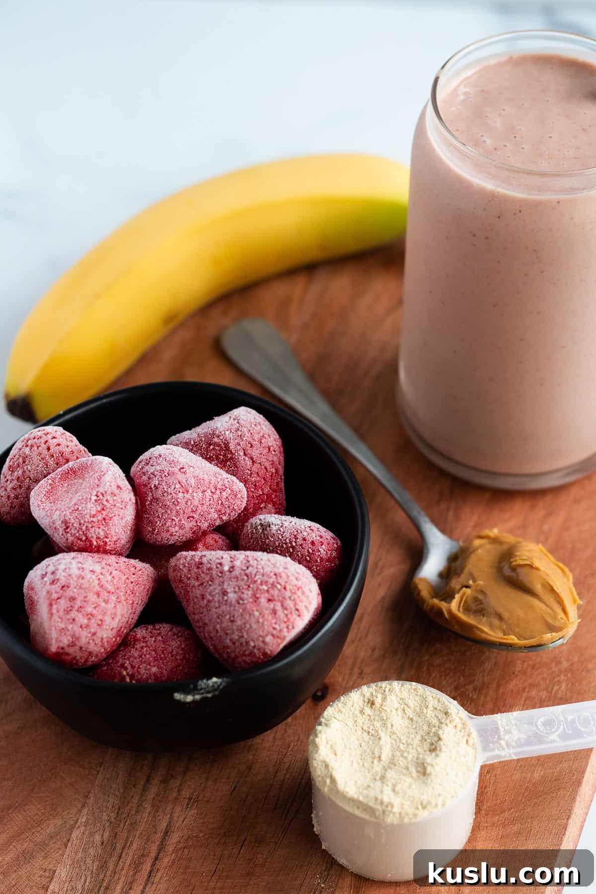 A wooden board with a smoothie, banana, frozen strawberries, protein powder and peanut butter.