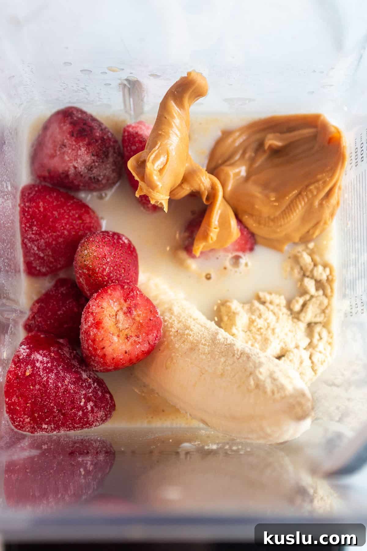 A blender jar with oat milk, a whole banana, frozen strawberries, peanut butter, and protein powder.