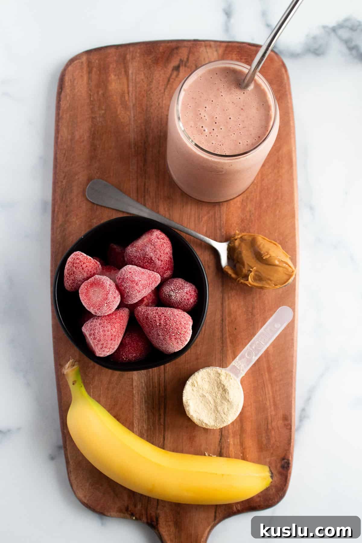 A wooden board with a glass filled with a smoothie, banana, bowl of frozen strawberries, peanut butter, and protein powder.