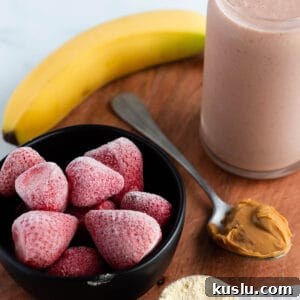 A wooden board with a glass filled with a pink smoothie, banana, frozen strawberries, scoop of peanut butter and protein powder.