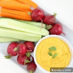 Quick 5-Minute Carrot Hummus Recipe for Busy Weeknights 5 Carrot hummus in a white ramekin with raw veggies on a white rectangular plate.