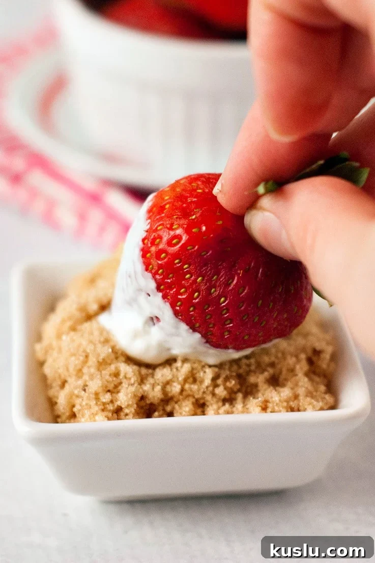 A fresh strawberry being rolled in brown sugar after dipping in sour cream