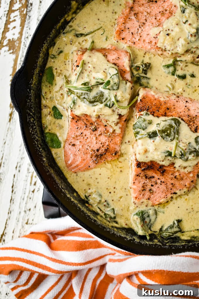 Three pan-seared salmon fillets sit in a creamy mustard and spinach sauce in a cast-iron skillet. A orange and white striped towel is around the handle.
